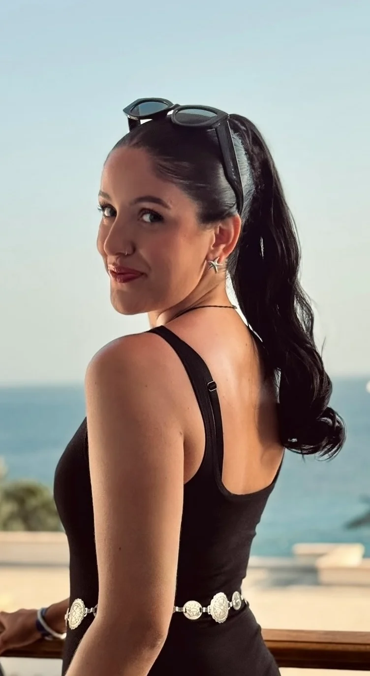 Woman with long dark ponytail, black top, and sunglasses on head, smiling with the sea behind her.
